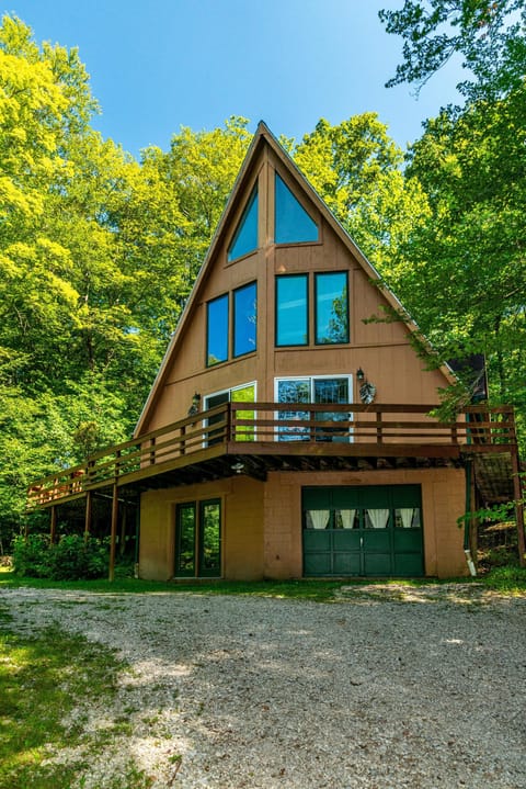 Exterior view of A-frame cottage with large wraparound deck, green garage door, and surrounding leafy trees.