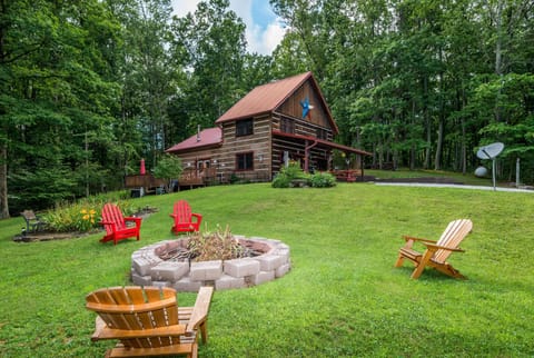Stone fire pit encircled by mixed wood and red Adirondack chairs on a grassy lawn in front of a log cabin.