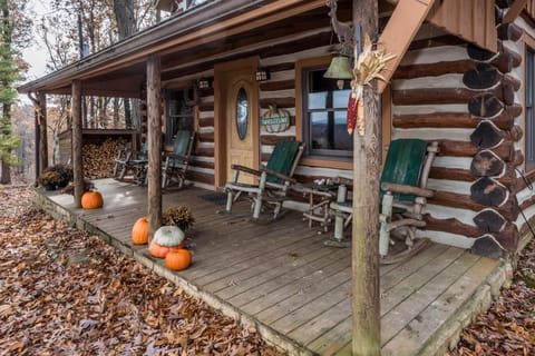 Wooden porch with rustic green rocking chairs, a stack of firewood, and autumn pumpkins scattered on fallen leaves.