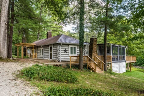 Exterior view of the 1930s log cabin with screened porch, stone chimneys, wooden deck, and surrounding trees.