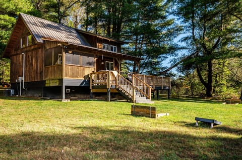 Rustic cabin exterior with a metal roof, screened porch, wooden deck, and grassy yard featuring cornhole boards.
