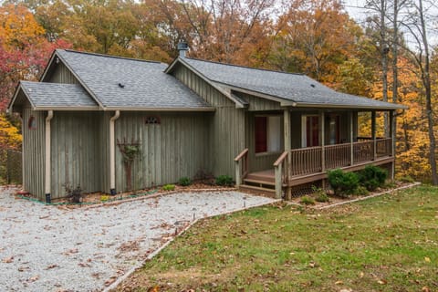 Exterior view of green wood-paneled cottage with covered porch, gravel driveway, and autumn trees surrounding.