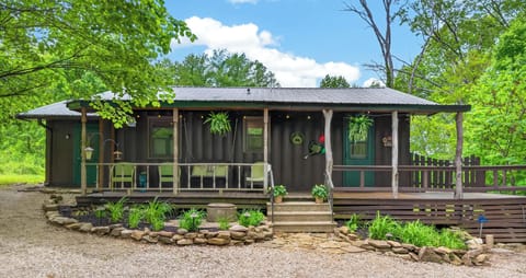 Gravel driveway and stone-edged garden bed lead to the cabin’s covered wooden porch with hanging ferns.