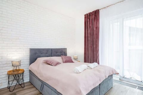 A bright and cozy studio featuring a large bed with light gray and pink bedding. A soft area rug and bedside tables with modern lamps complement the decor, with natural light streaming through a window with sheer curtains.