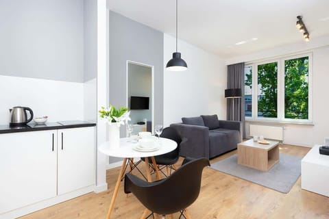 A bright living room with a sofa and coffee table, connected to a modern kitchen with white cabinets and built-in appliances. Large windows let in plenty of natural light.