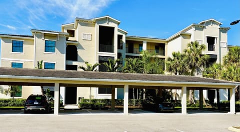 Covered parking spaces with beige stucco building and tropical landscaping in background under blue sky.