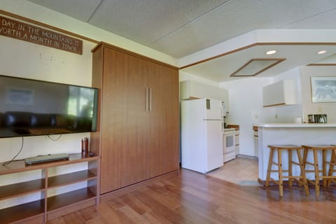 A small kitchen with wooden flooring, white appliances, bar stools, and a wall-mounted TV. A sign above the TV reads, "A day in the mountains is worth a month in town.