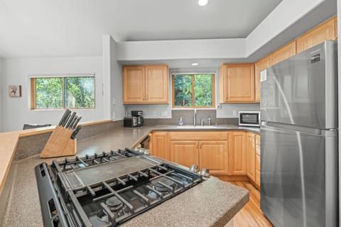 Spacious kitchen boasting natural wood cabinetry, a stainless steel refrigerator, and a sleek gas cooktop. Sunlight streams through large windows, creating a warm and inviting space to cook and gather after a day exploring the mountains.