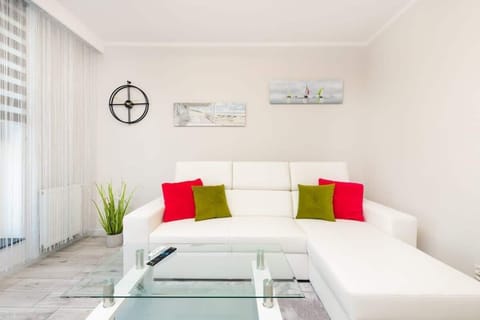  bright living room featuring a white sectional sofa with red and green accent pillows. A modern coffee table is positioned in front of the sofa, and there’s a small plant adding a touch of greenery. The room has large windows with blinds, allowing p