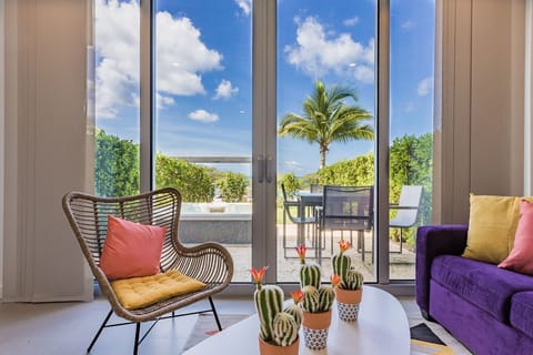 Curl up in your stylish living room as sunlight streams through floor-to-ceiling glass doors, framing vibrant tropical greenery and distant water views beyond your private patio.