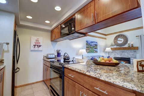 A modern kitchen with granite countertops, wooden cabinets, a black stove, microwave, and refrigerator. There is a fruit bowl on the counter and a painting and various decorative items on the walls.