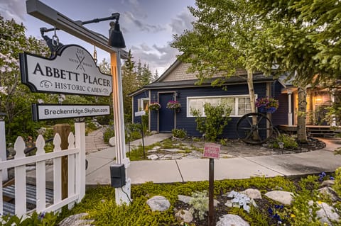 Image of the Abbett Placer Historic Cabin in Breckenridge. The dark blue cabin is surrounded by trees and greenery, with a pathway and a wooden sign at the entrance.