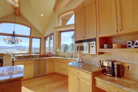 A modern kitchen featuring wooden cabinets, granite countertops, stainless steel appliances, and large windows with a scenic view. There are various kitchen items like a coffee maker and mugs on the counter.