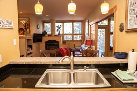 A view from the kitchen of a cozy living area with large windows, a fireplace, media setup, and dining table. Three pendant lights hang above the countertop and sink in the foreground.