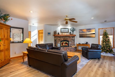 A cozy living room with leather sofas, a wooden coffee table, a lit fireplace, a Christmas tree, and a ceiling fan.
