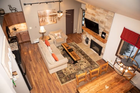 A cozy living area with a sofa, coffee table, stone fireplace, wall-mounted TV, and dining table. The space features hardwood floors, a kitchen, and a large window with a red valance.