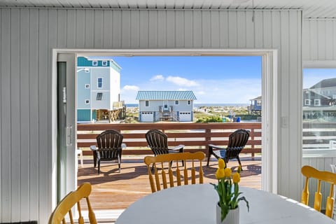 Dining area with a round white table, yellow chairs, sliding doors, deck seating, and a bright ocean view beyond neighboring beach homes.
