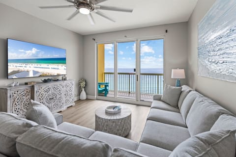 Relax in your coastal living room with ocean views through sliding glass doors, plush gray seating, and beachfront ambiance on the big screen.