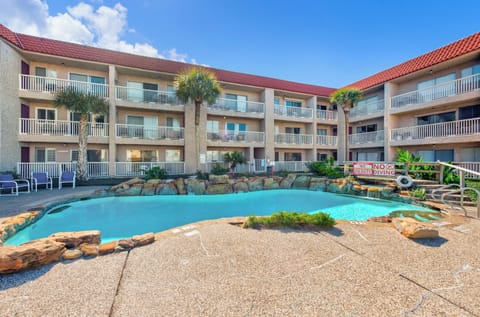 Multi-story property surrounds a sparkling pool with natural rock features and tropical palms under bright blue skies.