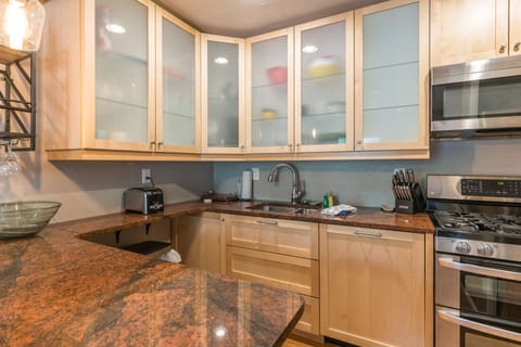 A modern kitchen featuring light wood cabinets with frosted glass doors, granite countertops, stainless steel appliances, and a sink with a pull-down faucet. A toaster and knife block are also visible.