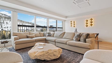 A bright, inviting living room where you'll relax on a spacious sectional sofa featuring cozy faux-fur pillows, taking in stunning snowy mountain views through floor-to-ceiling windows.