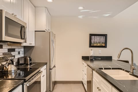 Modern kitchen with stainless steel appliances, black countertops, white cabinets, and an island with a sink. A framed picture of a forest is hung on the light-colored wall.