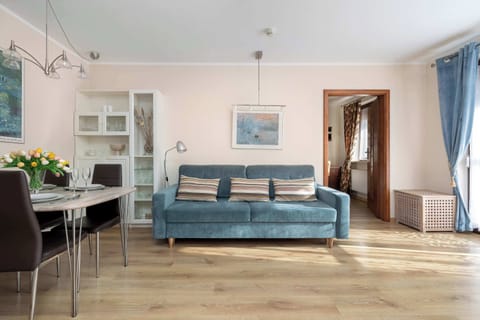 A living room with a blue sofa and armchairs is arranged around a small coffee table. Decorative artwork and shelves are mounted on the wall. The room appears spacious and well lit.