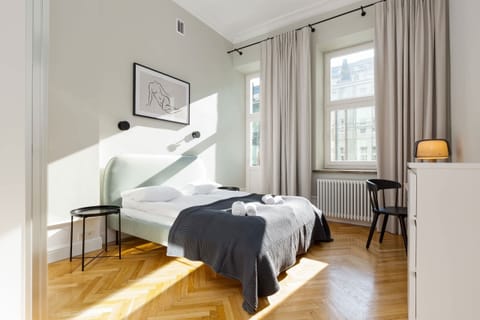 A spacious bedroom with a large bed, white linens, gray blanket, and elegant curtains letting in natural light.