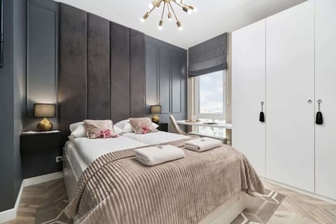 A bedroom with a plush double bed featuring beige and white bedding. A modern gray accent wall with soft lighting adds a luxurious touch.