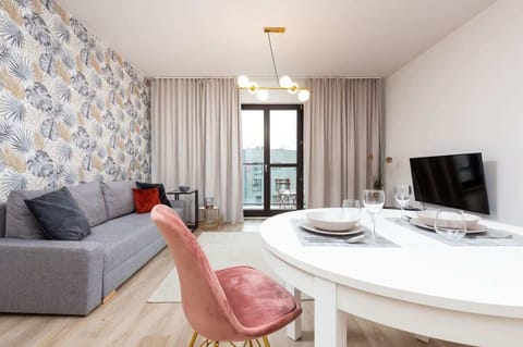 Bright living room with grey sofa against floral patterned wallpaper. In the foreground is a table with a round top and a pink chair.