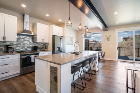 Modern kitchen with white cabinets, stainless steel appliances, a gray tile backsplash, and a large island with four bar stools. Sliding glass doors open to a scenic mountain view.
