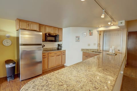 A kitchen with granite countertops, wooden cabinets, a stainless steel refrigerator, microwave, and coffee maker. A wall clock and trash bin are also visible.