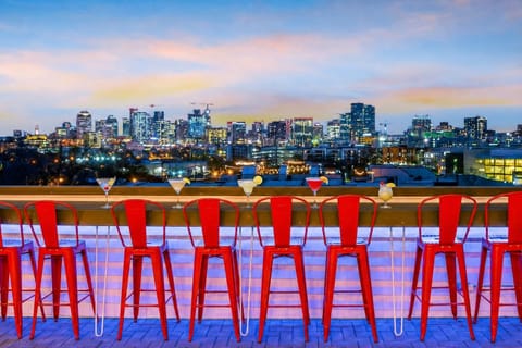 Red bar stools line the rooftop edge, the downtown skyline beyond, twilight settling in. Twelve of you fit here, everyone in the conversation