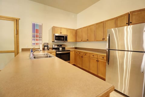 A modern kitchen with wooden cabinets, stainless steel appliances, a double sink, and a large countertop. A colorful wall art piece and a coffee maker are visible by the microwave.