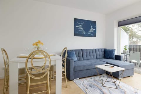 A bright and cozy living room with a blue sofa, a round white dining table paired with elegant chairs, and a large window letting in natural light, decorated with minimalist wall art.