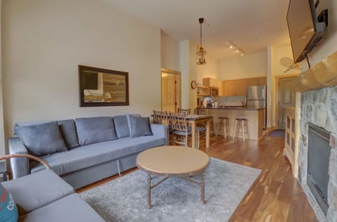 A living room with a grey couch, wooden coffee table, fireplace, wall-mounted TV, and a view into a bright kitchen area with a bar counter and stools.