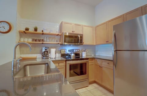 A modern kitchen with stainless steel appliances, wooden cabinets, a sink, and an electric stove. Shelves above the counter hold dishes and plants. A round clock is on the wall.