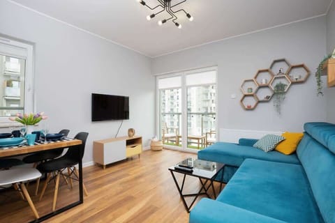 An open-plan living room with a blue sofa, a dining table set for two, and bright natural light flooding in through large windows.