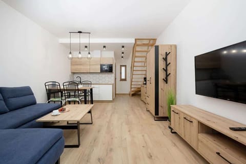 Another angle of the living room showing the dining area in the background. The layout is open-plan, with a small dining table and chairs near the back, maintaining a natural, wooden aesthetic throughout.
