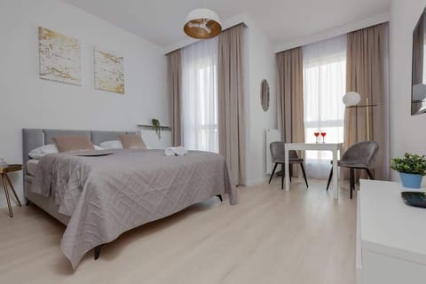 A spacious, modern studio with soft beige tones, featuring a large bed, a seating area with a table, and ample natural light coming through large windows.