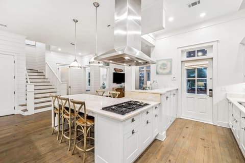 Cook and gather in this pristine white kitchen featuring a spacious island with built-in gas range, elegant pendant lighting, and warm wood floors.