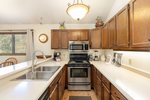A tidy kitchen with wooden cabinets, white countertops, stainless steel appliances, and a double sink. A dining area is visible through a pass-through window on the left.