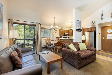A cozy living room and kitchen area featuring a sofa, an armchair, a wooden coffee table, a dining table with chairs, a modern kitchen, and large windows with a view of greenery.