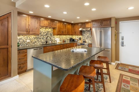 Modern kitchen featuring a central island with four wooden stools, wooden cabinets, a tiled backsplash, a stainless steel refrigerator, and various kitchen appliances.