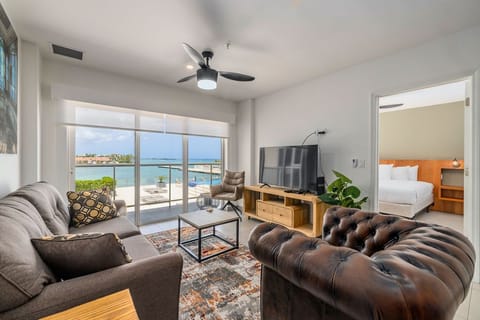 Unwind in your stylish living area with stunning turquoise water views through floor-to-ceiling windows. A coastal breeze flows through as you relax in plush seating.