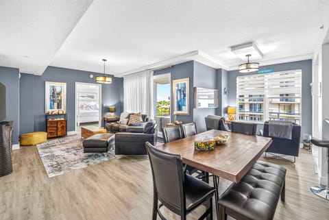 Spacious open-plan living and dining area with sleek leather furnishings and warm wood tones. Sunlight streams through large windows, creating a welcoming atmosphere perfect for relaxing.