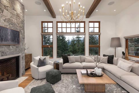 - Cozy mountain living room with tall windows overlooking forest views  
- Stone fireplace and chandelier lighting create warm alpine ambiance  
- Plush sectional seating perfect for après-ski relaxation  
