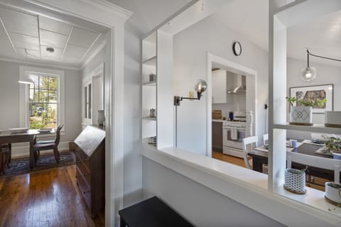 Bright, open kitchen and dining area with white walls, wooden floors, modern light fixtures, and a view into a sunlit room with a table and chairs.