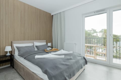 A cozy bedroom featuring a double bed with gray bedding and a wooden accent wall behind it. Large windows offer natural light and a view of greenery.