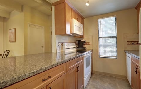 A kitchen with wood cabinets, granite countertops, a white oven and microwave, tile backsplash, and a large window with blinds.